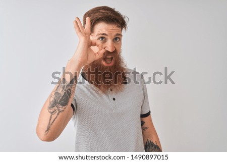 Young redhead irish man wearing grey polo standing over isolated white background smiling positive doing ok sign with hand and fingers. Successful expression.