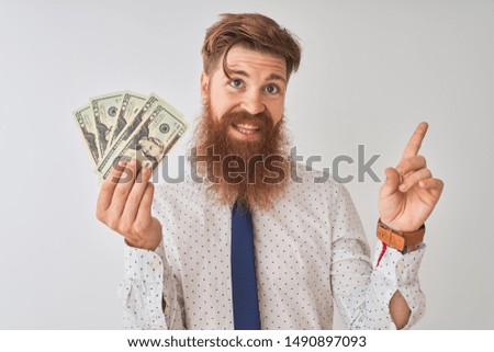Young redhead irish businessman holding dollars standing over isolated white background very happy pointing with hand and finger to the side