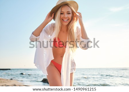 Young pretty cheerful blond woman in swimsuit wearing white shirt and hat happily looking in camera with sea on background