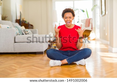 Young beautiful african american woman sitting on the floor at home shouting with crazy expression doing rock symbol with hands up. Music star. Heavy concept.
