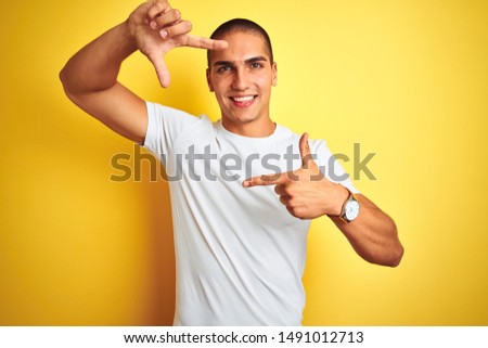 Young caucasian man wearing casual white t-shirt over yellow isolated background smiling making frame with hands and fingers with happy face. Creativity and photography concept.