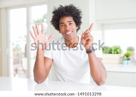 Young african american man wearing casual white t-shirt sitting at home showing and pointing up with fingers number six while smiling confident and happy.