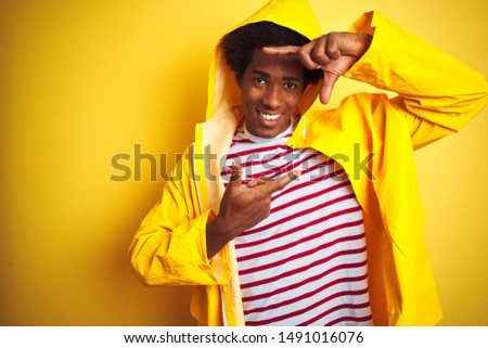 African man with afro hair wearing rain coat with hood standing over isolated yellow background smiling making frame with hands and fingers with happy face. Creativity and photography concept.