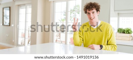 Wide angle shot of young handsome man at home smiling with happy face winking at the camera doing victory sign. Number two.