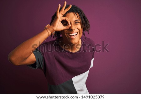 Afro man with dreadlocks wearing casual t-shirt standing over isolated purple background doing ok gesture with hand smiling, eye looking through fingers with happy face.