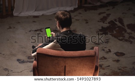 Attractive young man with brown hair sitting in comfortable chair with white pillow and sliding on iPhone green screen in dark room. Stock footage. Chroma key