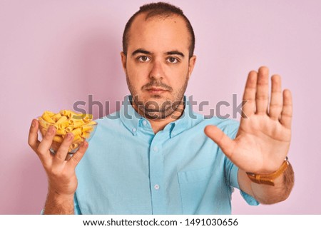 Young man holding bowl with macaroni pasta standing over isolated pink background with open hand doing stop sign with serious and confident expression, defense gesture