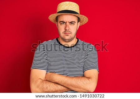 Young man wearing striped navy t-shirt and hat standing over isolated red background skeptic and nervous, disapproving expression on face with crossed arms. Negative person.