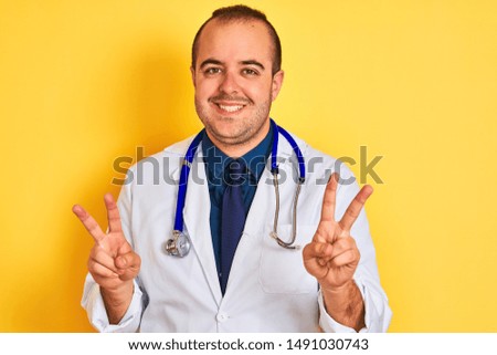 Young doctor man wearing coat and stethoscope standing over isolated yellow background smiling looking to the camera showing fingers doing victory sign. Number two.