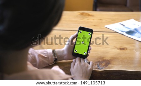 View from the shoulder of woman with brown hair in pink shirt holding iPhone in hand and looking at the chroma key green screen. Stock footage. Cellphone with chroma key screen