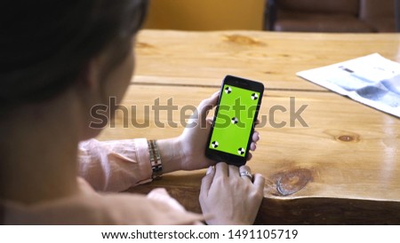 Young woman with brown hair in pink shirt sitting at the wooden table, holding iPhone in hand and looking at the chroma key green screen. Stock footage. Smartphone new technology concept