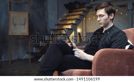 Close-up of young handsome man in black shirt and jeans looking at digital tablet while sitting in big comfortable chair at home. Stock footage. Modern decorated home interior on the background