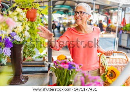 Portrait of a beautiful senior woman doing shopping in a market on a sunny morning, she carries a basket with fruits and vegetables and a bouquet of flowers. Woman buying flowers on the local market

