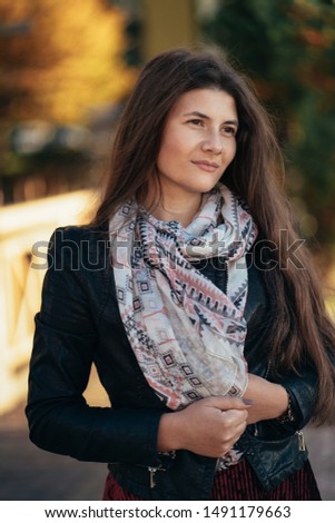 A young beautiful brunette girl with long hair in a leather jacket and a scarf laughing on a sunny summer day. Female standing on city street. Fashion Look. Fashionable young woman outdoor