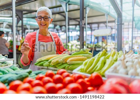 Friendly woman tending an organic vegetable stall at a farmer's market and selling fresh vegetables from the garden. Female Stall Holder At Farmers Fresh Food Market