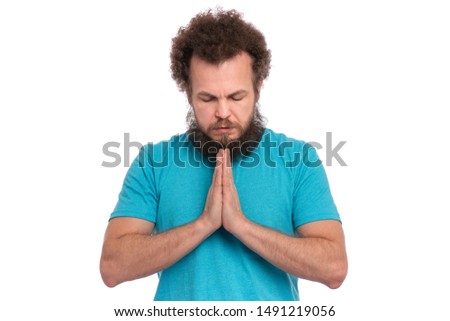 Faithful bearded handsome man stands with keeps hands together and closes eyes. Spirituality and calm atmosphere concept. Religious man praying to God with folded hands, isolated on white background