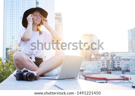young girl photographer sitting with camera and laptop outdoors and retouching photo, tourist freelancer on a background of city sunset