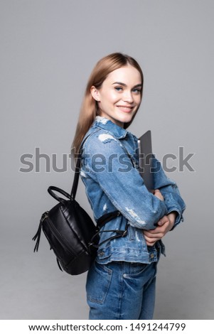 Indoor photo of college student girl isolated on gray background, smiling at camera, pressing laptop to chest, wearing backpack, ready to go to studies, start new project and suggest new ideas.