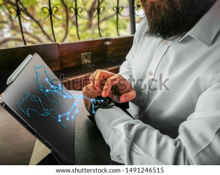 bearded adult rich businessman touching economy touching his smart watch and connecting to his pro tablet and internet world map holographic