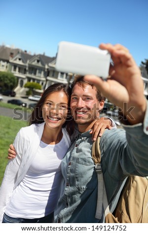 Couple fun taking self-portrait photo with cell phone camera. Multiracial happy tourist couple on travel vacation in San Francisco, Alamo Square, USA.