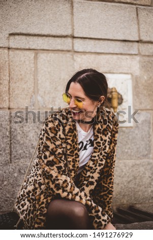 Lovely young laughing brunette woman posing outdoor on old stone fountain background. Wearing stylish yellow glasses, trendy leopard print faux fur coat.