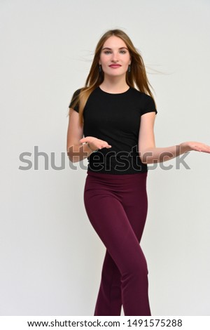 Full-length vertical portrait of a pretty brunette girl with long flowing hair in a black t-shirt and burgundy trousers on a white background. Smiling, showing emotions.