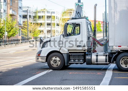 Day cab big rig white professional semi truck with roof spoiler transporting commercial cargo in dry van semi trailer running on the urban city street with multilevel buildings Royalty-Free Stock Photo #1491585551
