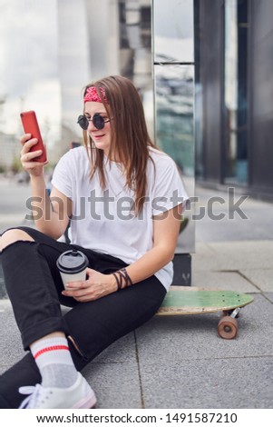 Photo of brunette in sunglasses with phone in her hands sitting on skateboard on background of modern buildings in city