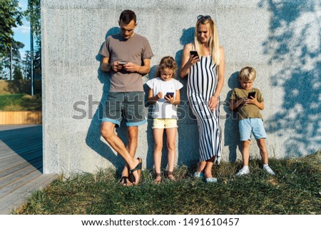 Picture of blonde woman and man and children with phones in their hands standing on concrete wall on street