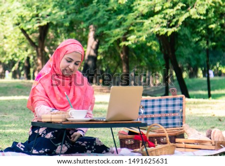 Lifestyle Happy beautiful Arabian Muslim woman in hijab sitting in the park working with laptop and breakfast or writing checklist on paper notebook, lifestyle working Muslim woman technology concept
