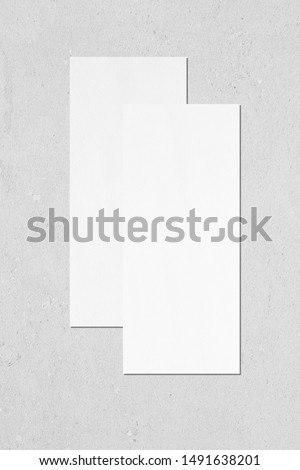 Two empty white vertical rectangle price-list or menu mockups with soft shadows lying on top of each other on neutral light grey concrete wall background. Flat lay, top view