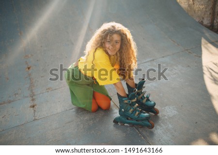 Young woman girl playing in green and yellow clothes and orange stockings with curly hairstyle with rollers on hand. Roller skating female in skate park.