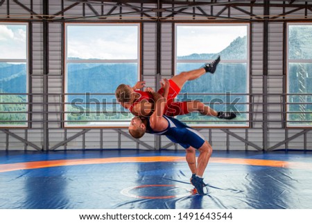 Two men in blue and red wrestling tights are wrestlng and making a suplex wrestling on a  blue wrestling carpet in the gym on the background of mountains. The concept of male wrestling and resistance