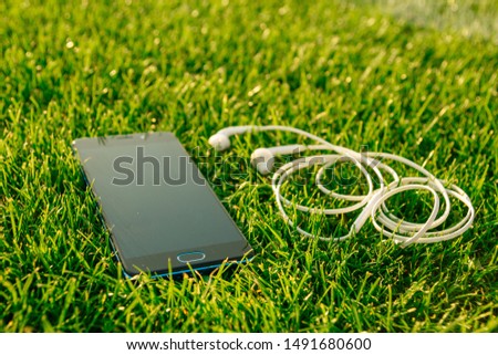 Smartphone with white headphones lies on the green grass. 