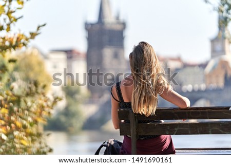 Young attractive female traveler sit down on a wooden bench with a backpack in front of a blurred background with the historical Bridge of Charles in Prague. 