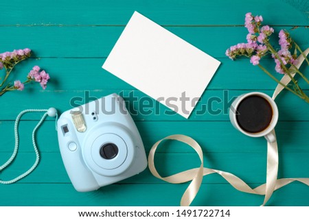 Modern instant camera, coffee cup, blank paper card, flowers on green wooden background. Top view, minimal flat lay style composition. Women's desk, fashion blogger, beauty technology