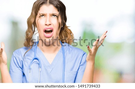 Young adult doctor woman wearing medical uniform crazy and mad shouting and yelling with aggressive expression and arms raised. Frustration concept.