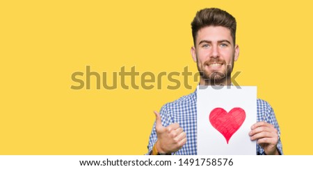 Young handsome man holding paper with red heart happy with big smile doing ok sign, thumb up with fingers, excellent sign