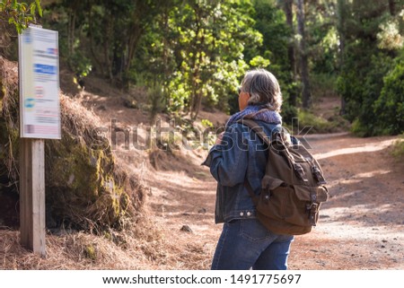 Rear view of a senior woman with gray hairs and brown backpack. Looking at panel to choose the best excursion in the woods. Casual clothing  for excursion. Enjoying and protect the nature