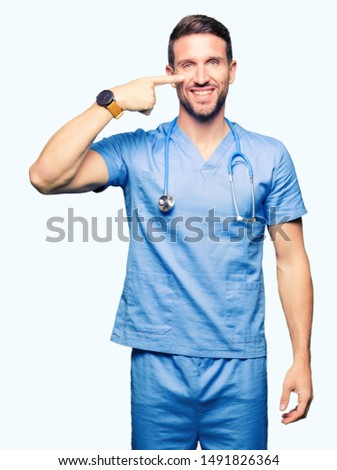 Handsome doctor man wearing medical uniform over isolated background Pointing with hand finger to face and nose, smiling cheerful