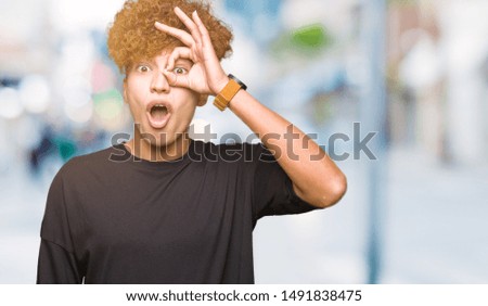 Young handsome man with afro hair wearing black t-shirt doing ok gesture shocked with surprised face, eye looking through fingers. Unbelieving expression.