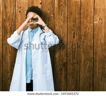 Young african american doctor woman wearing medical coat over isolated background Doing heart shape with hand and fingers smiling looking through sign