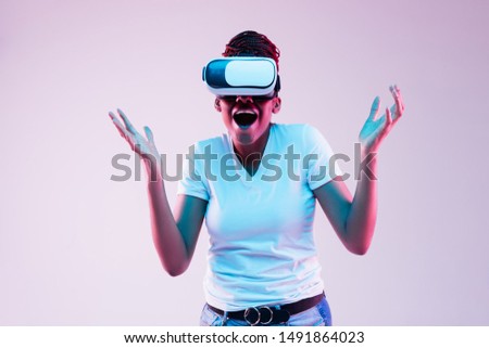 Portrait of young african-american woman's playing in VR-glasses in neon light on gradient background. Concept of human emotions, facial expression, modern gadgets and technologies. Look astonished.