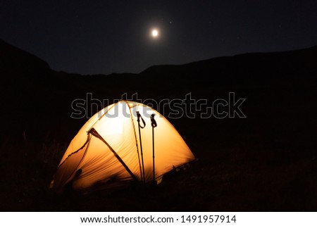 The camping tent is lit by a lantern at night in the mountains. Bright moon in the sky. Mountain climbing hike.
