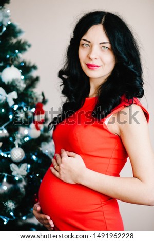 Closeup photo of pregnant woman posing near Christmas tree at home. Merry Christmas and Happy Holidays! Pregnancy, holidays, people and expectation concept.