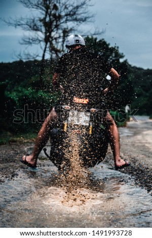 Man wearing black and white helmet is riding the motorcycle through the deep puddle on the tropical island after the rain. Drops of water and pieces of dust are raising up from under the wheels.