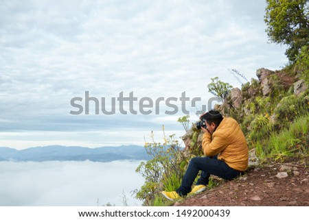 close up on a man sitting on top of a mountain during a cloudy and foggy day underneath, taking pictures with his professional camera towards some blue mountains