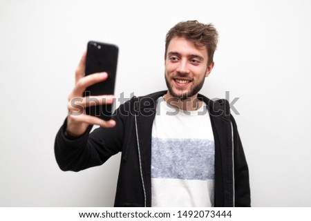 Portrait of positive, comic, trendy guy with stubble shooting selfie on smart phone, using gadget device isolated on gray.