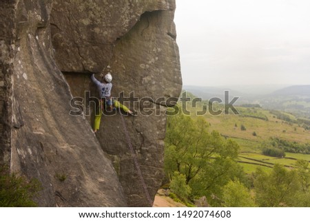 Young man climbing on a wall with beautiful english countryside in the background. View of a professional male climber with green trousers from behind.
