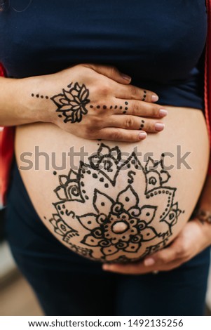 Master is drawing henna tatto on pregnant belly, closeup. A mehendi artist paints a beautiful mandala. Motherhood concept, happy maternity